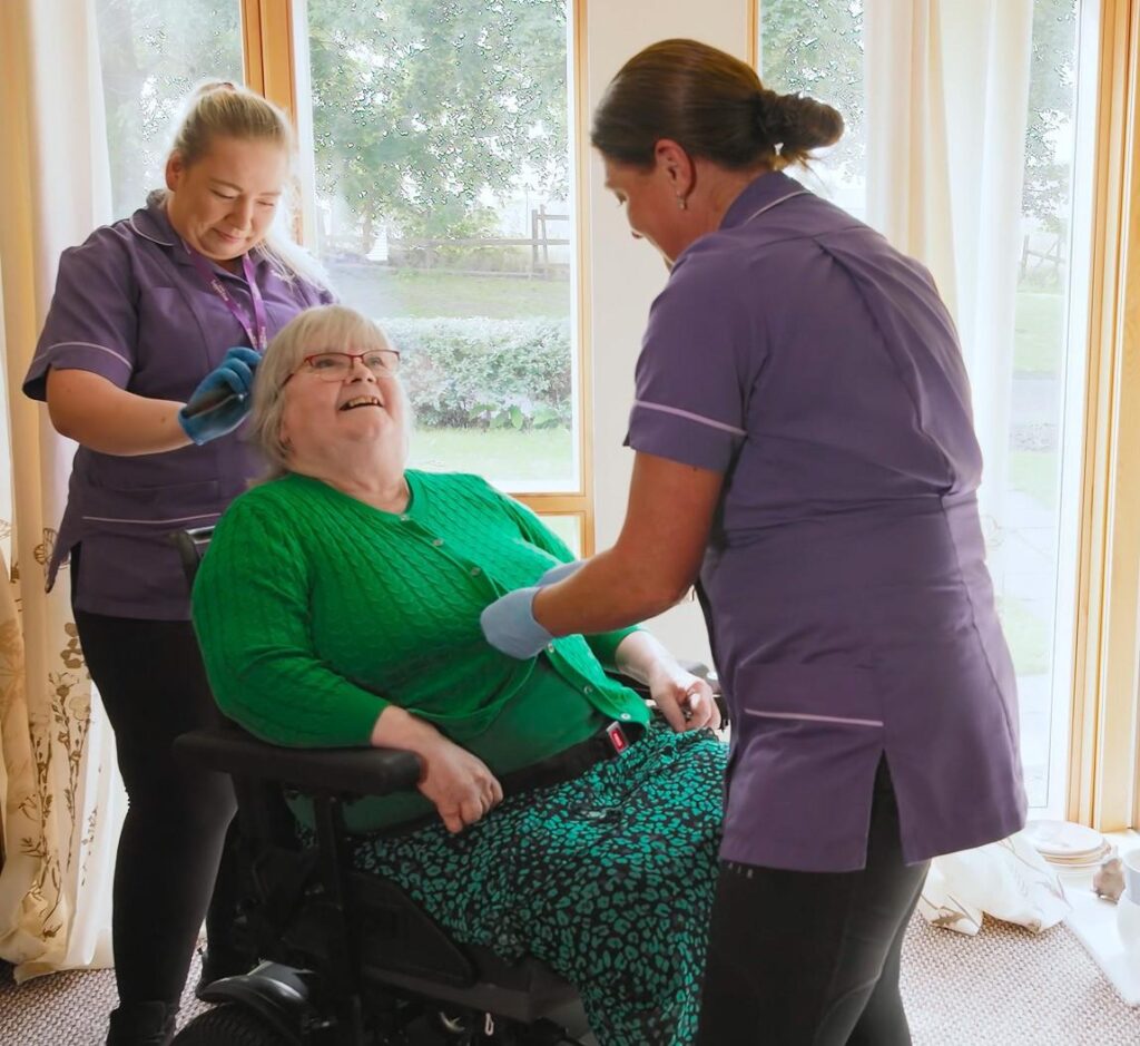 image of 3 Women, two women in healthcare purple uniforms are assisting a female patient in a transport chair